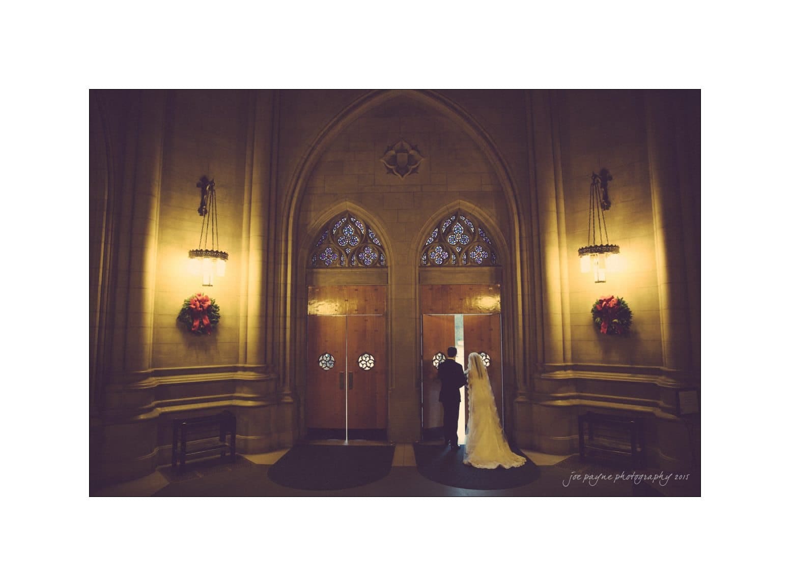 A bride and groom stand inside a large, ornate church entrance with tall wooden doors and intricate stained glass. The space is dimly lit, creating a warm, intimate atmosphere. Red wreaths adorn the walls on either side of the doorway.