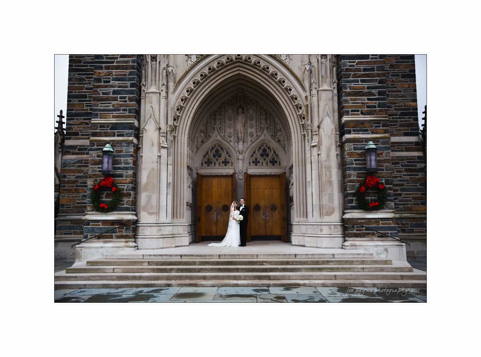 A bride and groom share a kiss in front of a large, ornate stone church entrance. The doors are wooden and flanked by Christmas wreaths with red bows. The architecture features intricate stone carvings and arches.