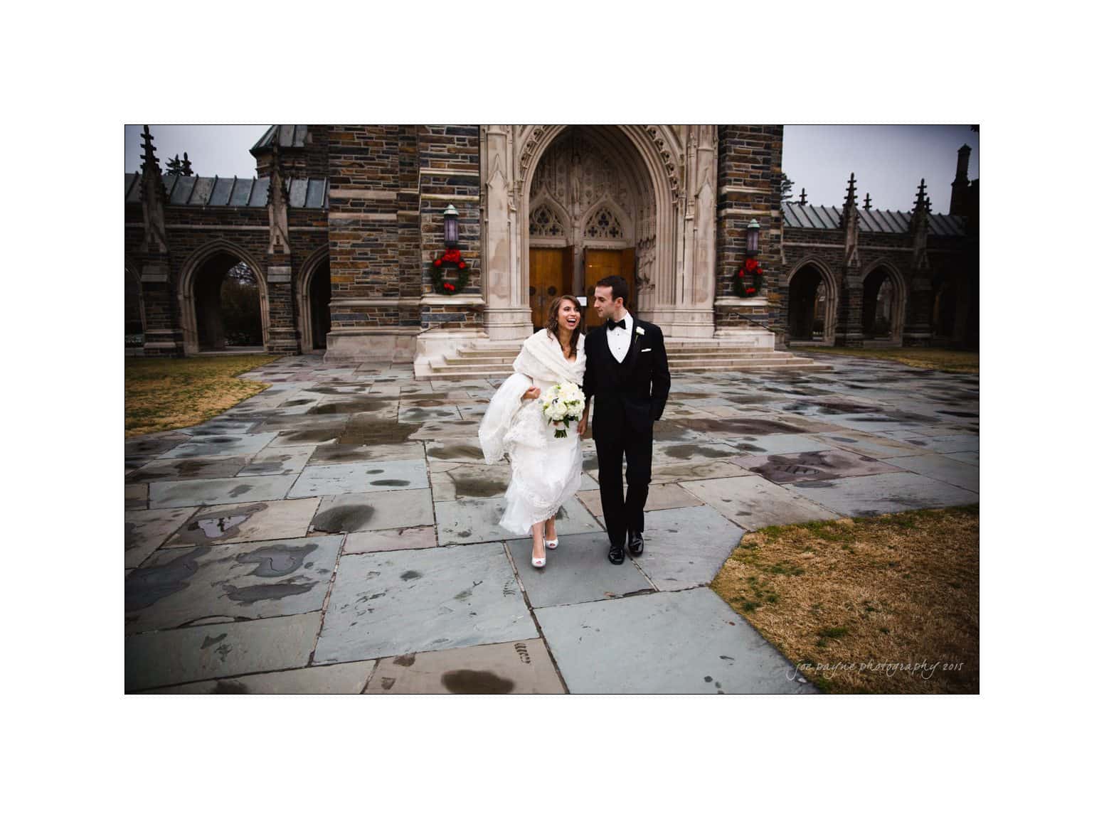 A bride and groom walk arm in arm, smiling at each other outside a stone cathedral. The bride wears a white dress, carries a bouquet, and is wrapped in a shawl. The groom is in a black tuxedo. The ground is wet, suggesting recent rain.