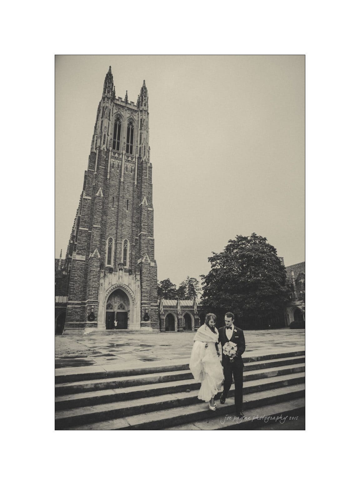 A black and white photo of a couple in wedding attire walking down steps, with a tall, ornate Gothic-style tower in the background. The groom holds a bouquet, and the sky is overcast, creating a classic, timeless atmosphere.