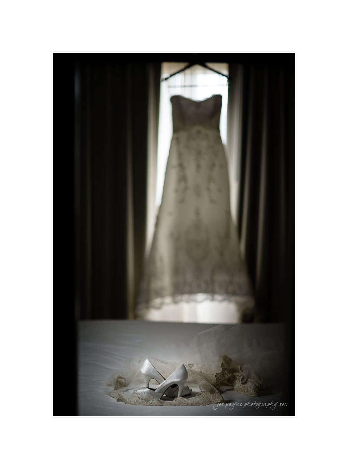 A white, intricately detailed wedding dress hangs against a softly lit window. In the foreground, white heeled shoes and a delicate veil are artfully arranged on a bed, creating a serene bridal scene.