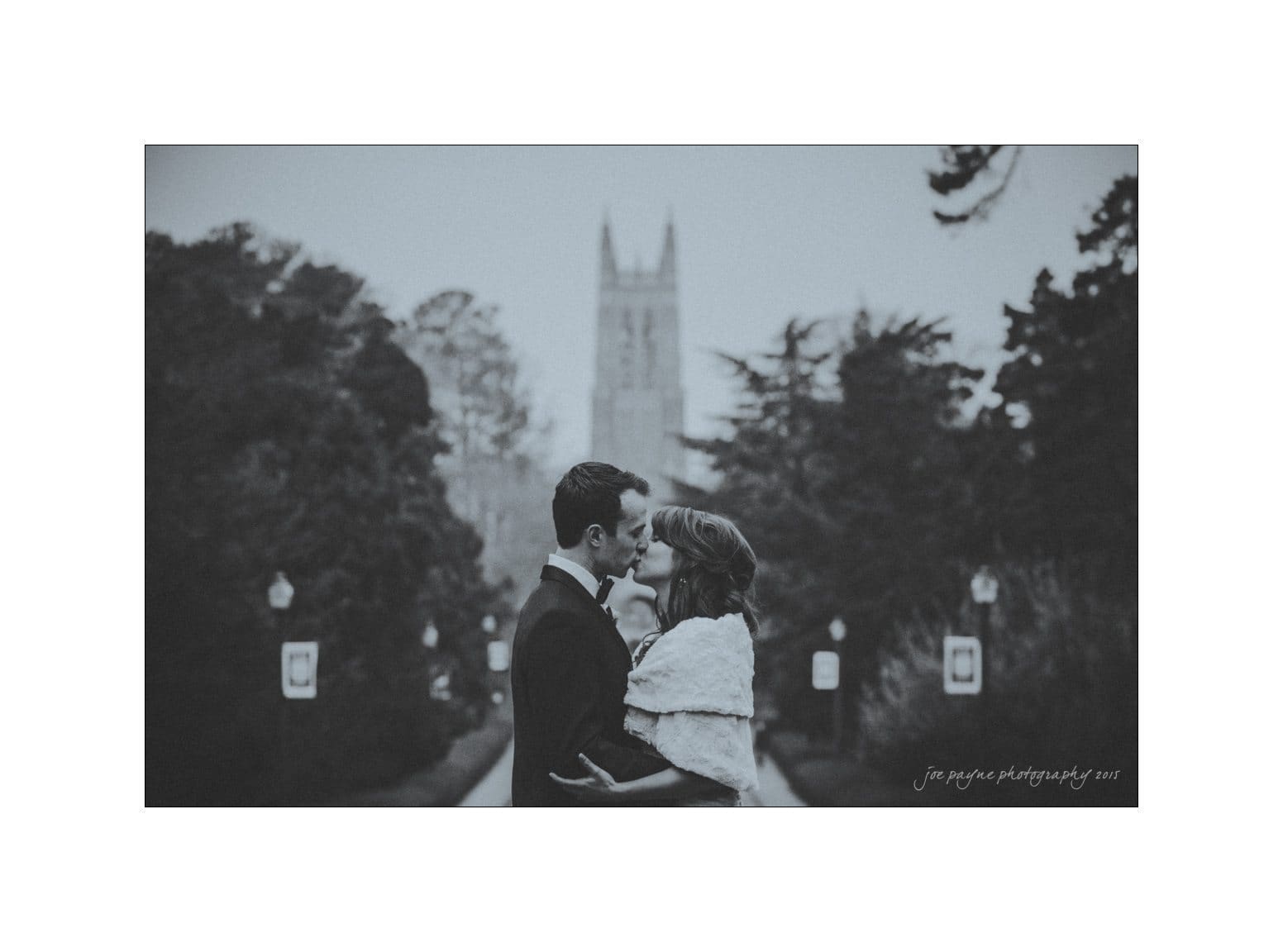 A black and white photo of a couple embracing and about to kiss in front of a blurred, distant cathedral. They stand on a path lined with trees, wearing formal attire. The atmosphere is romantic and timeless.