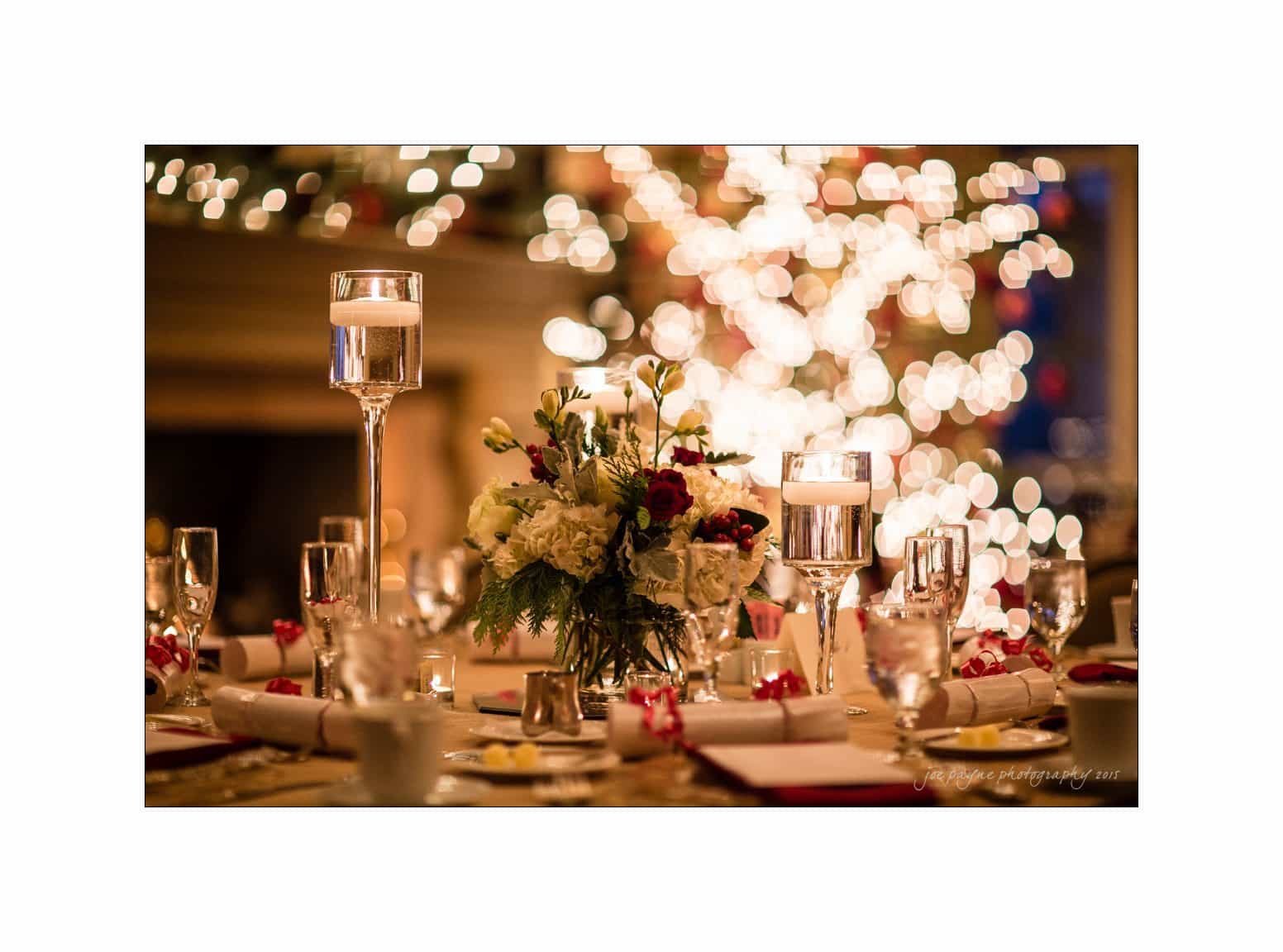A beautifully decorated table set for a festive event, featuring a centerpiece with white and red flowers, surrounded by glowing candles. The background is brightly illuminated with bokeh lights, creating a warm and inviting atmosphere.