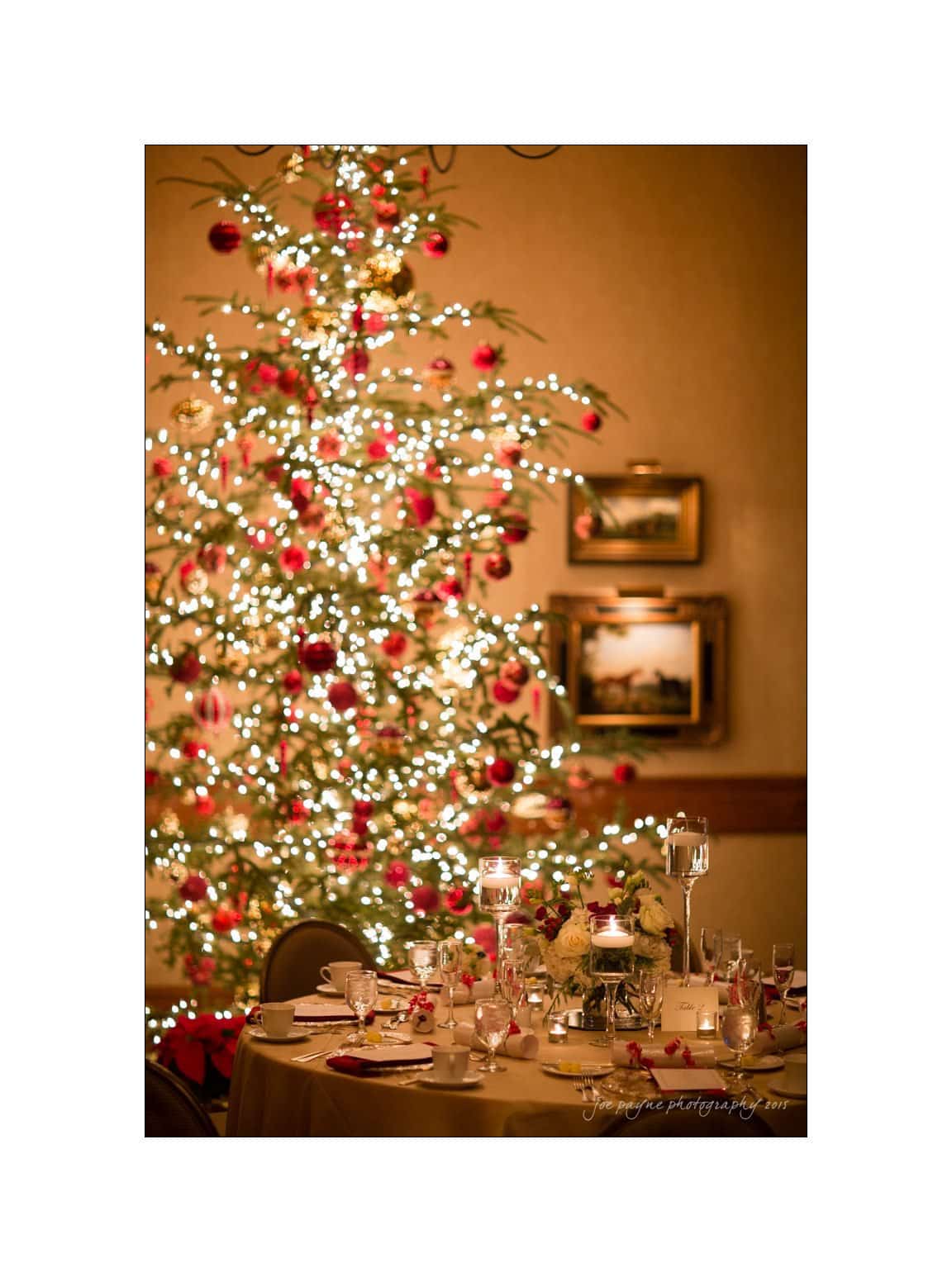 A beautifully decorated Christmas tree with glowing lights and red ornaments stands next to a round dining table set with elegant tableware, candles, and floral centerpieces in a warmly lit room.