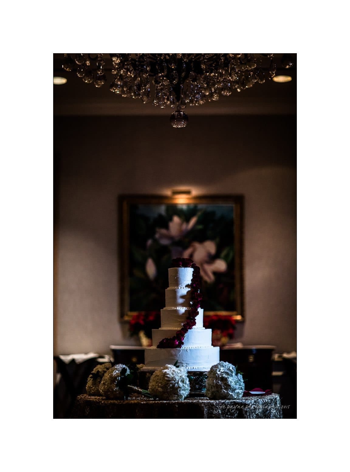 A five-tiered white wedding cake adorned with red flowers is set on a decorated table. The lighting highlights the cake against a dimly lit room, with a chandelier overhead and a floral painting in the background.