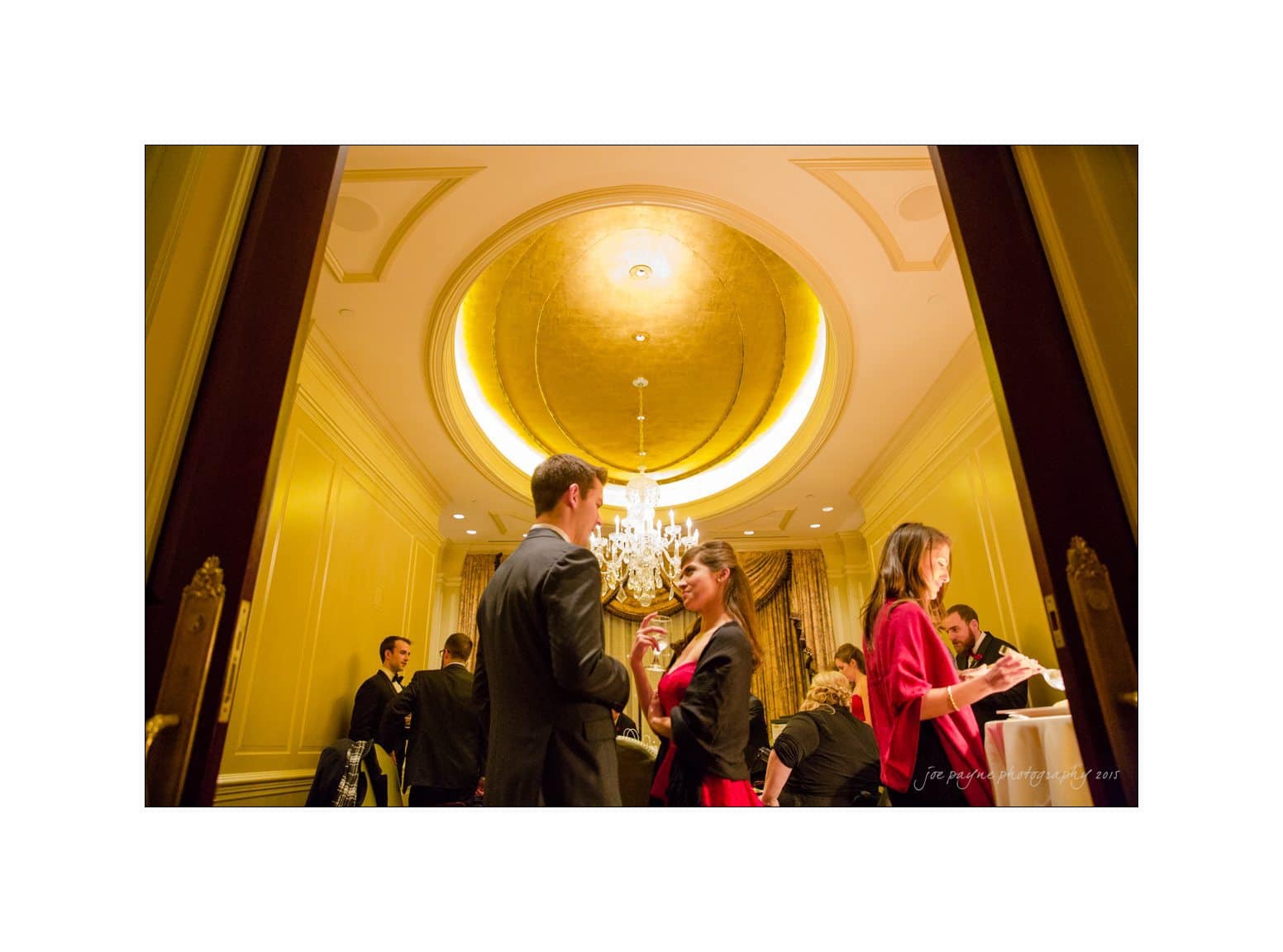 A formal gathering in an elegant room with a golden ceiling and chandelier. Guests in suits and dresses engage in conversation, while others stand near tables. The atmosphere is sophisticated and warm.
