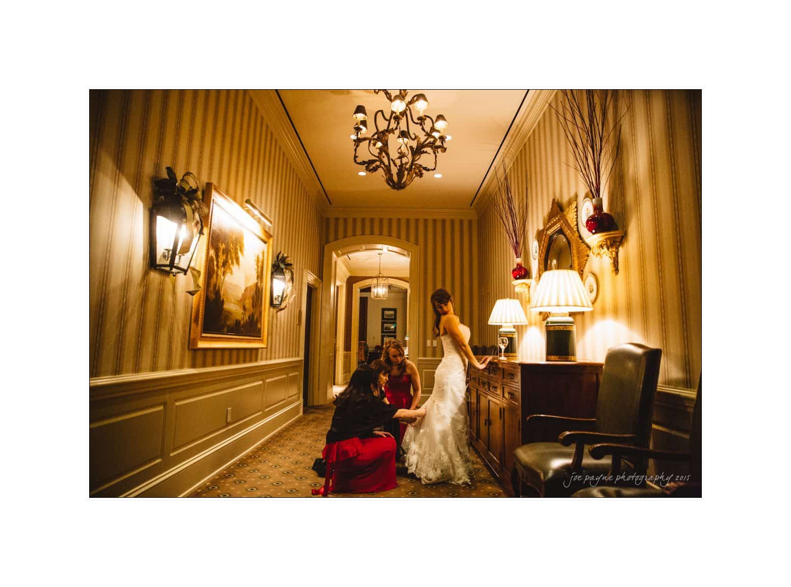 A bride in a white gown stands in a warmly lit hallway as two women in red dresses help adjust her dress. Elegant sconces and a chandelier add to the vintage ambiance of the room.