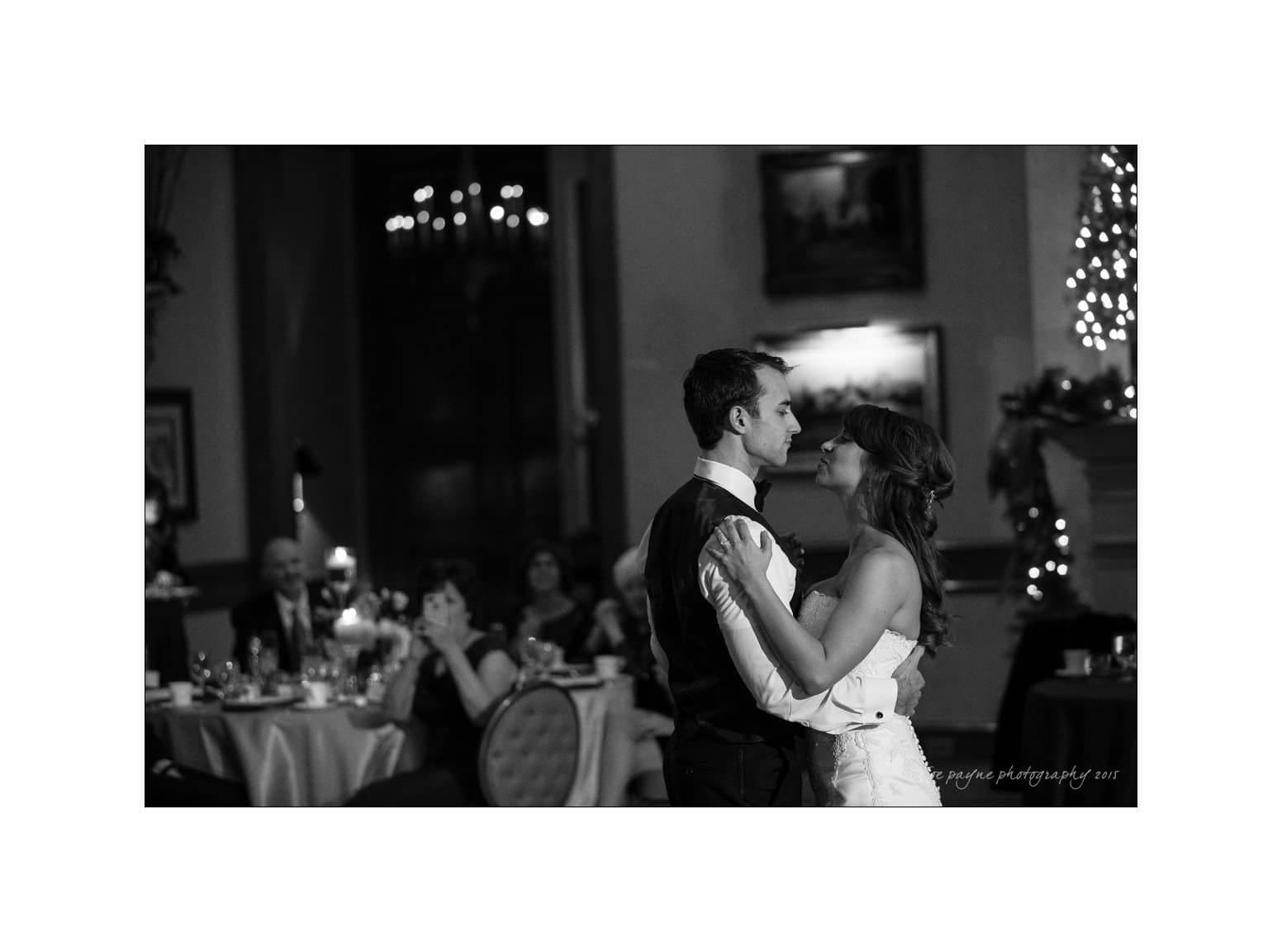 A bride and groom share their first dance in an elegantly decorated venue. Guests watch from their tables adorned with candles and decor, creating a romantic atmosphere. The room is softly lit, highlighting the couple in the center.