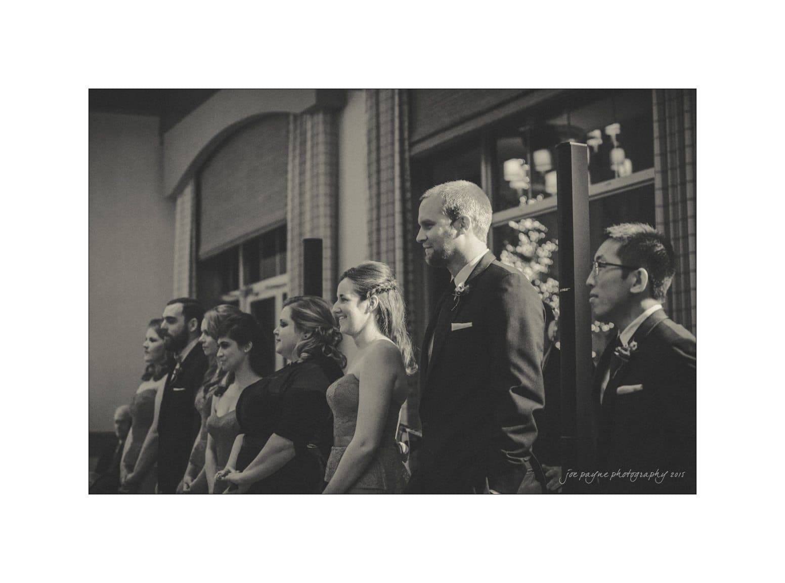 A black-and-white photo of a group of people standing in a line, dressed in formal attire, likely at a wedding or formal event. They appear attentive and are inside a building with large windows in the background.