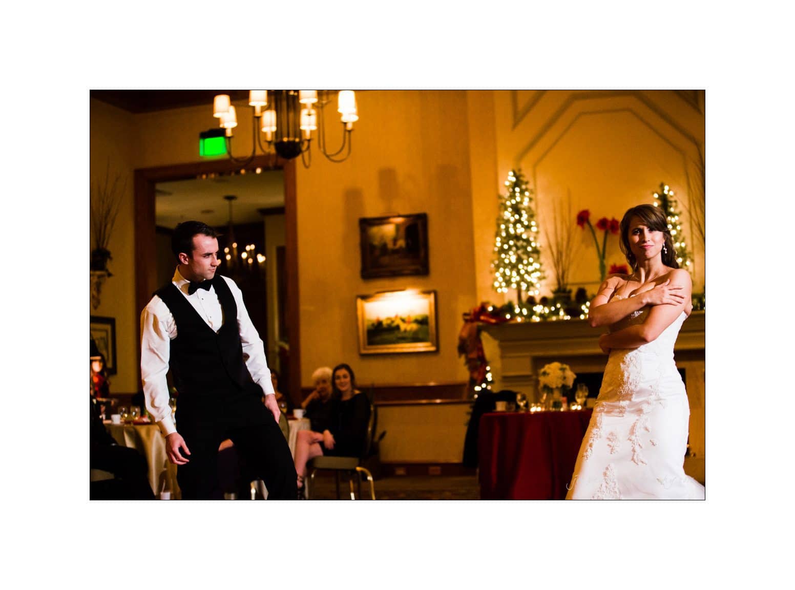 A bride in a white gown and a groom in a vest and bow tie perform a dance routine. The room is decorated with Christmas lights and a decorated tree in the background, with guests watching from their seats.