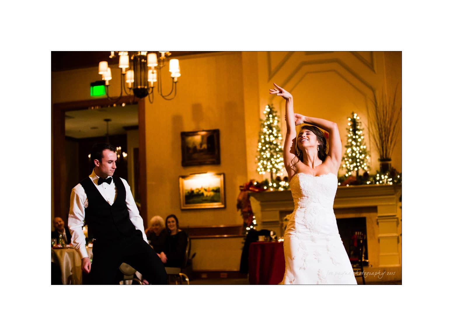 A bride and groom dance enthusiastically at a wedding reception. The bride wears a white strapless gown, and the groom is in a black and white tuxedo. They are in a warmly lit room with holiday decorations and guests seated in the background.