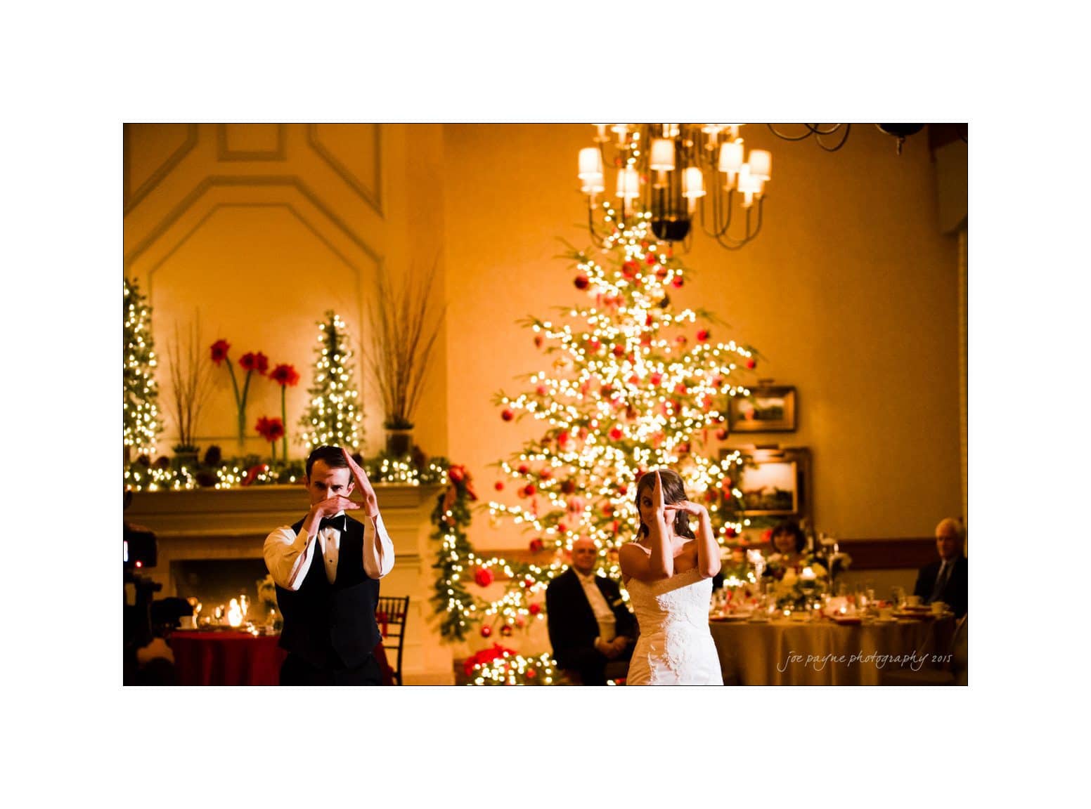 A bride and groom are dancing energetically in a warmly lit room decorated for Christmas. A large, illuminated Christmas tree and a fireplace with lights and red decorations serve as the backdrop. Guests are seated at tables in the background.