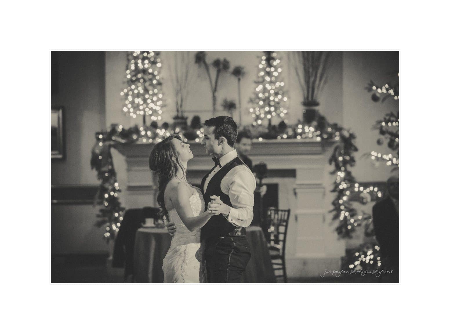 A couple dances together in a warmly lit room, decorated with festive lights and ornaments. They share a joyful moment in front of a fireplace adorned with garlands and small, lit Christmas trees. The scene is captured in black and white.