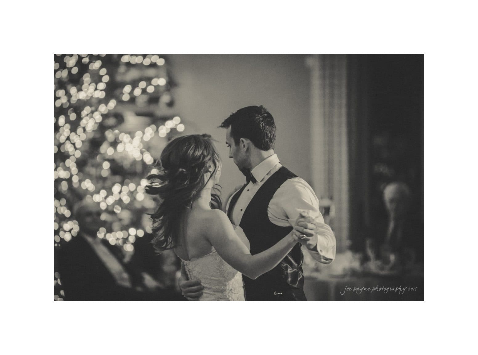 A bride and groom share a dance in a softly lit room. The bride is wearing a strapless gown, and the groom is in a vest and bowtie. A decorated, illuminated tree is visible in the background. The image is in black and white.