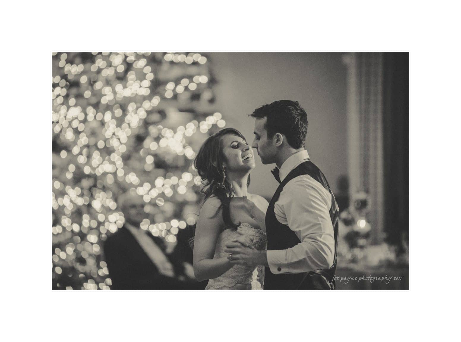 A couple shares a romantic first dance at their wedding, with the bride in a lace gown and the groom in formal attire. Soft, dreamy lighting creates a magical ambiance, with a beautifully lit Christmas tree in the background.