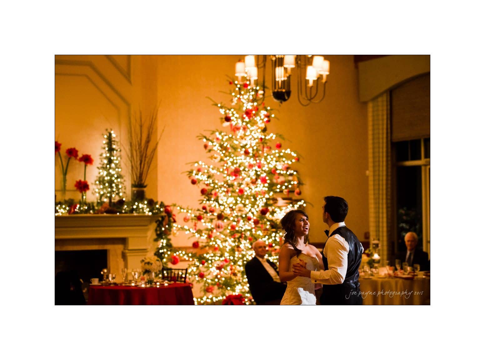 A couple dances in a warmly lit room, with a beautifully decorated Christmas tree in the background. Elegant chandeliers hang from the ceiling, and guests sit at tables enjoying the festive atmosphere.