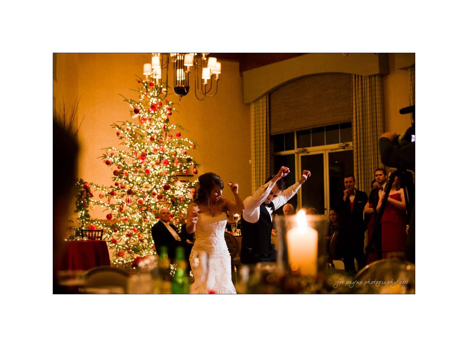 A bride and groom dance joyfully at a wedding reception in a warmly lit room with a large, decorated Christmas tree in the background. Guests watch and cheer as the couple celebrates.