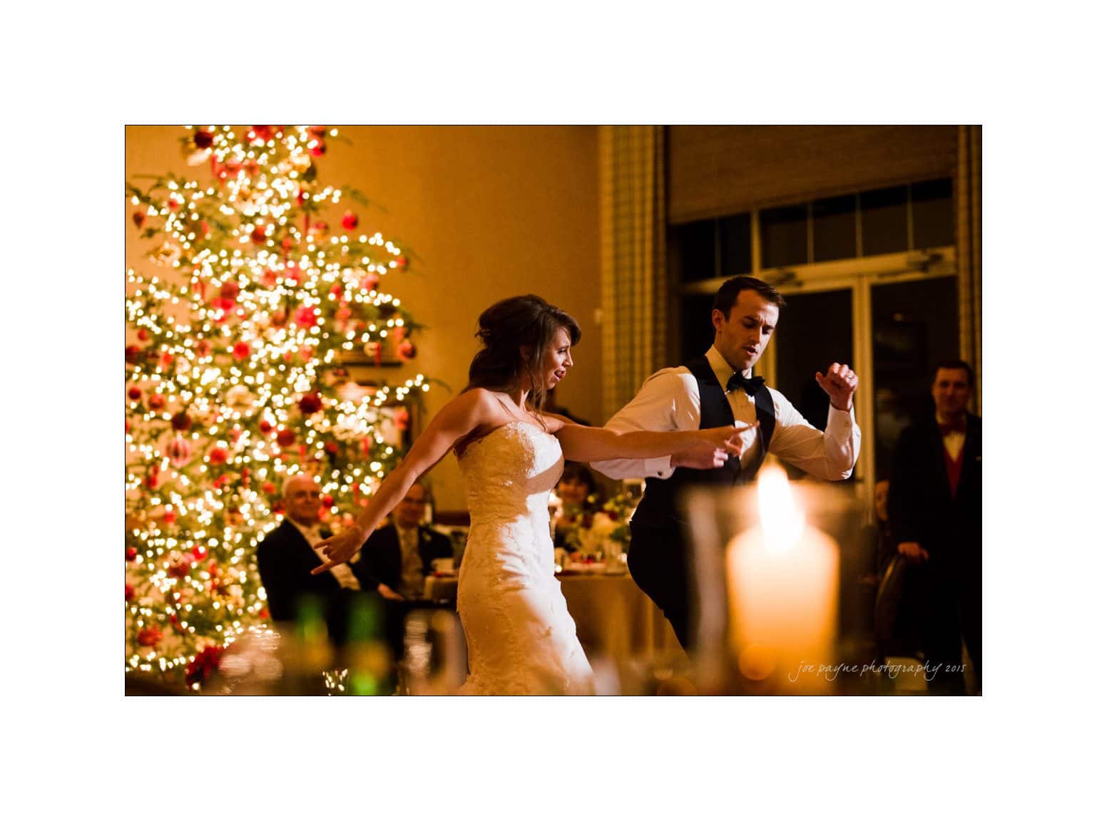 A bride and groom dancing energetically at their wedding reception. The bride wears a white gown, and the groom is in a vest and bow tie. A lit Christmas tree decorates the background, adding a festive glow to the scene.