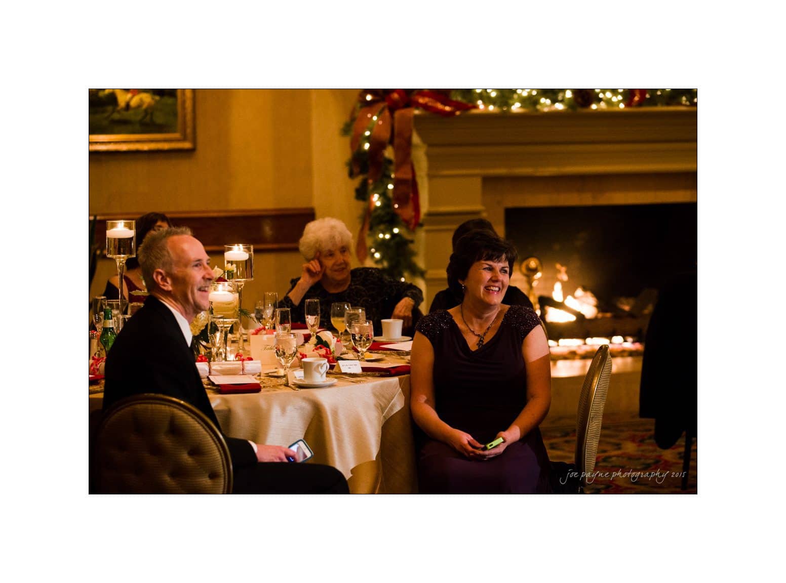People are seated at round tables decorated with candles, enjoying a festive gathering in a warmly lit room. A fireplace and Christmas decorations can be seen in the background.