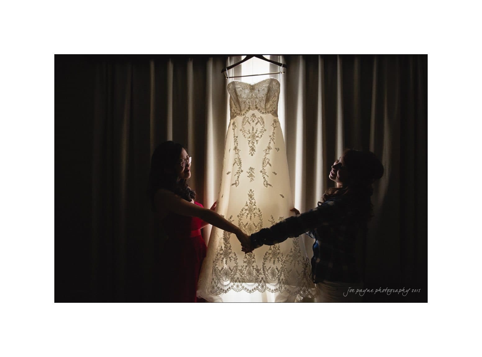 Two people holding hands in front of a window with a wedding dress hanging in the center. The lighting creates a silhouette effect against the curtains.
