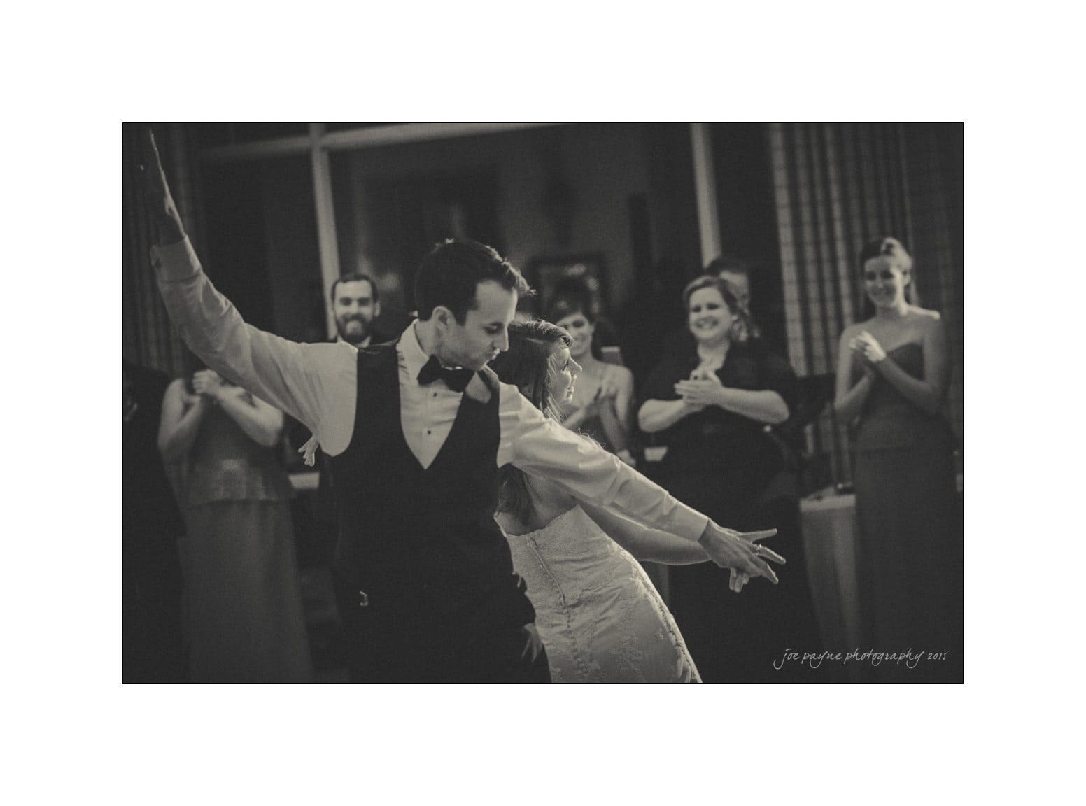 A couple in formal attire is joyfully dancing at a wedding reception. The groom is in a vest and bow tie, and the bride is in a lace dress. Guests in the background are clapping and smiling, creating a celebratory atmosphere.