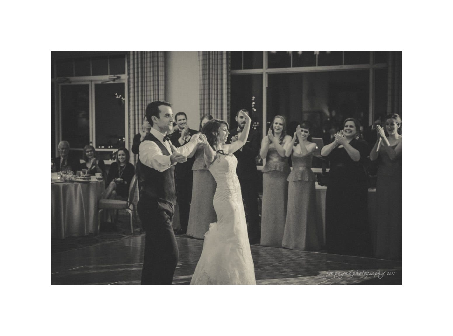 A couple dances joyfully at their wedding reception, surrounded by smiling and applauding guests seated at tables. The scene has a warm, celebratory atmosphere in a softly lit room.
