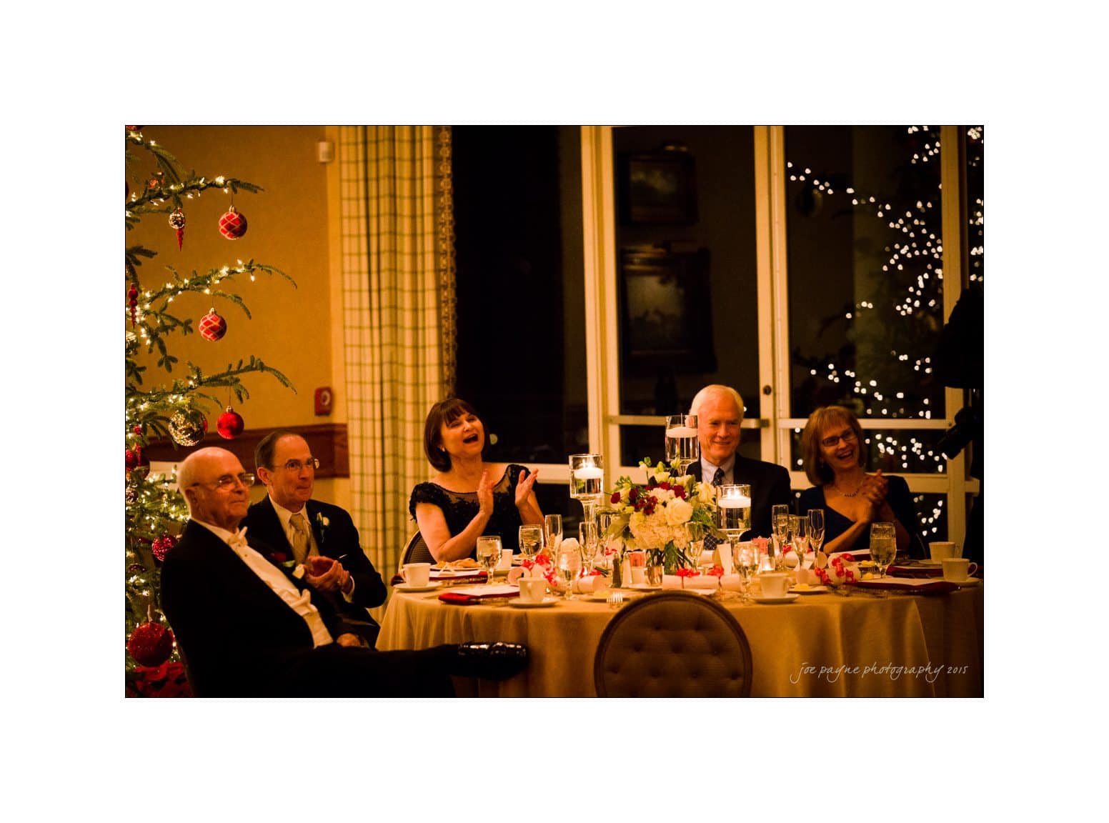 A group of elegantly dressed people sitting around a table decorated with candles and flowers at a festive event. There is a Christmas tree with red ornaments in the background, and twinkling lights are visible through the windows.