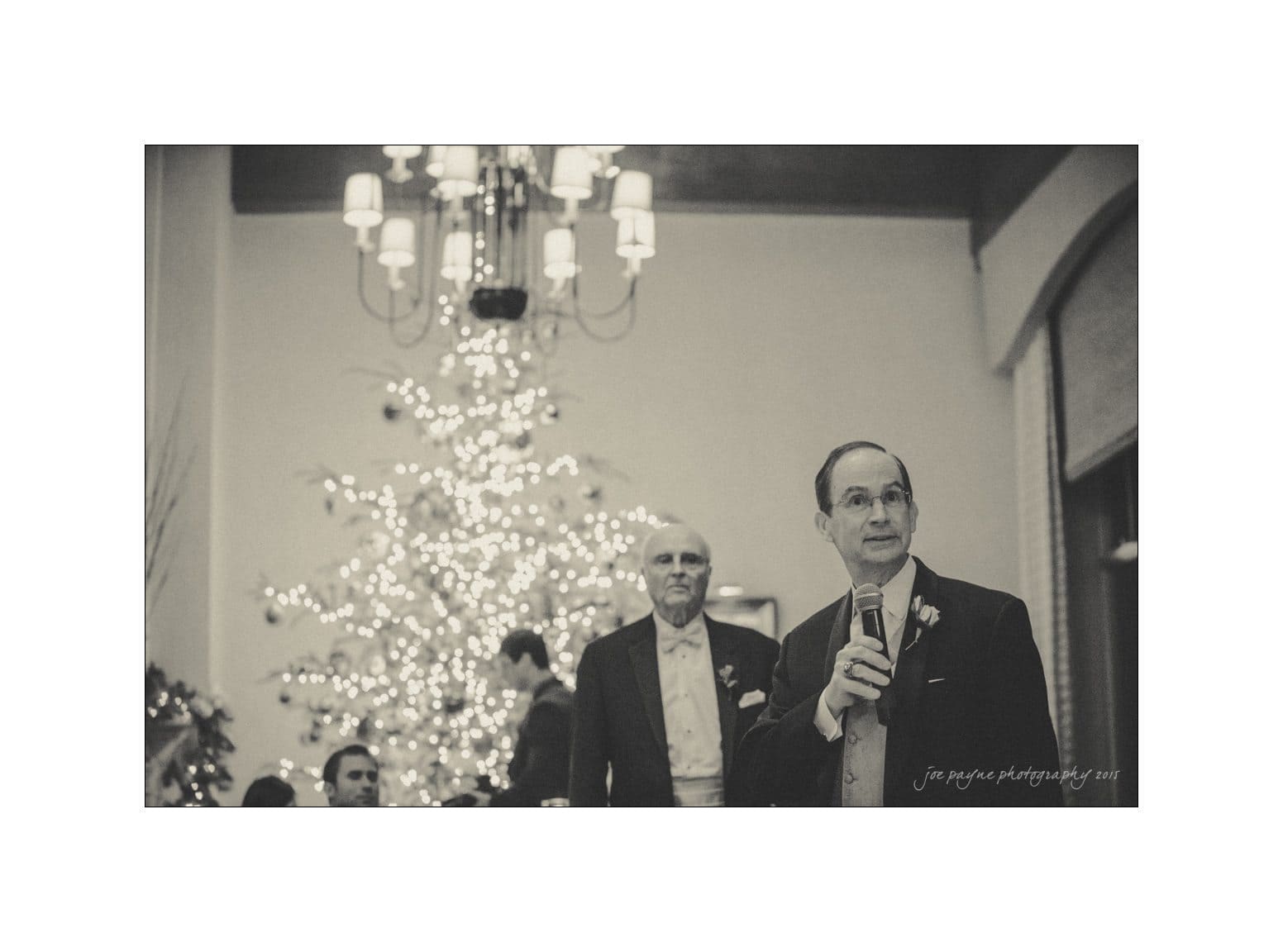 A man stands holding a microphone, appearing to speak at an event. Behind him, another man watches. The background features a large, decorated Christmas tree with glowing lights and an elaborate chandelier above. The scene is captured in a black and white tone.