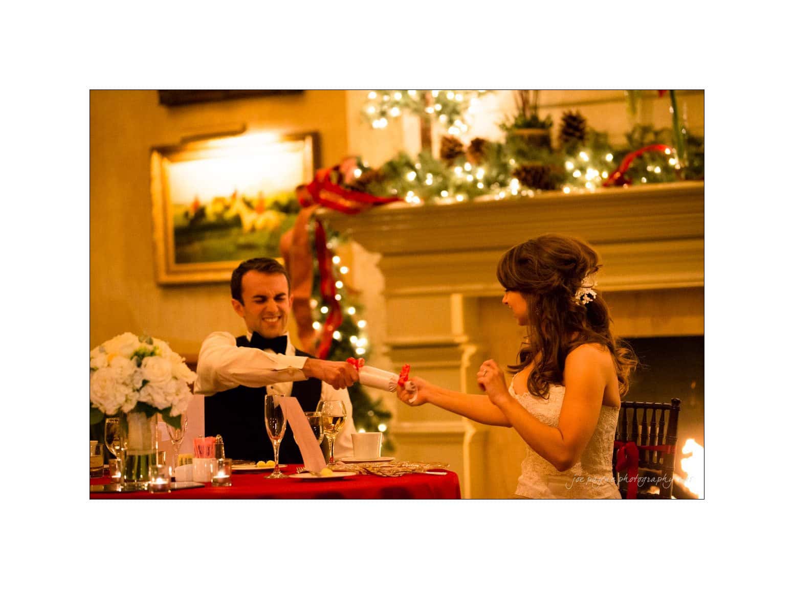 A couple at a festive dinner table pulls a Christmas cracker, surrounded by holiday decorations and soft lighting. The woman, in a white dress, and the man, in a suit, are smiling and engaged in the moment.