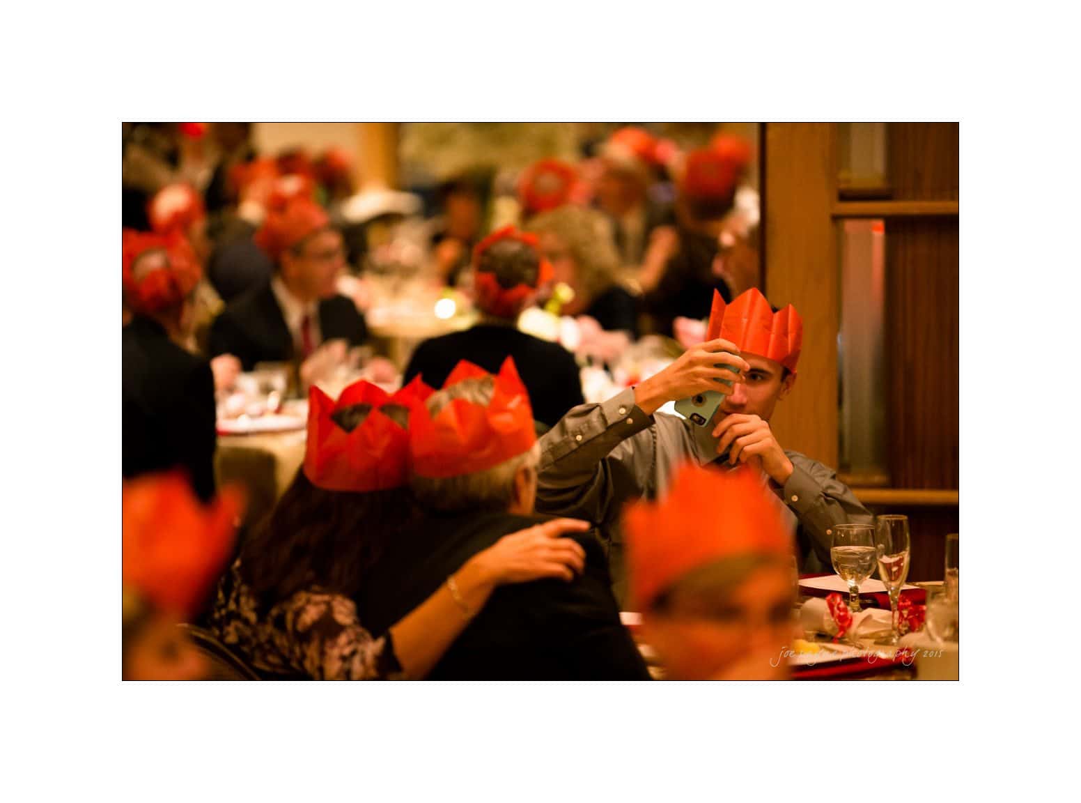 People wearing red paper crowns sit at a festive dinner, with one person taking a selfie. The scene is warm and celebratory, with tables set with wine glasses and soft lighting.