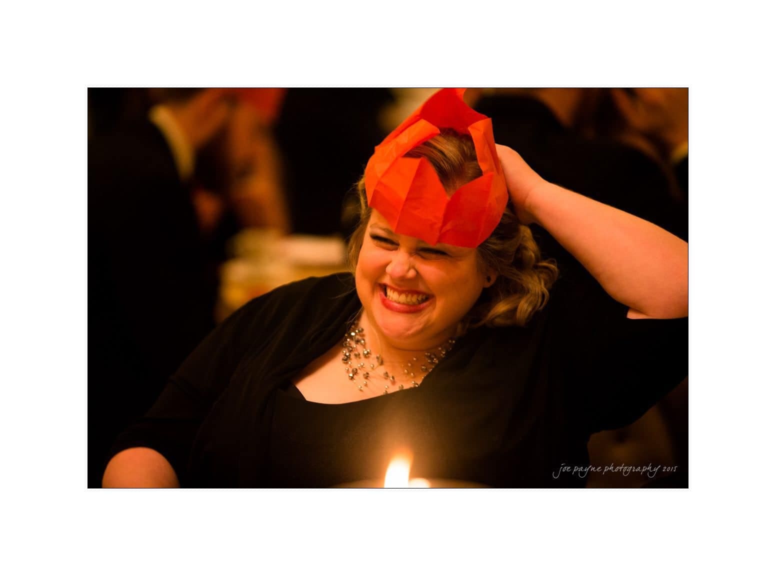 A person smiling and wearing a red paper crown while sitting at a dimly lit event. They are wearing a black outfit with a sparkling necklace. The background is blurred, with other people visible.
