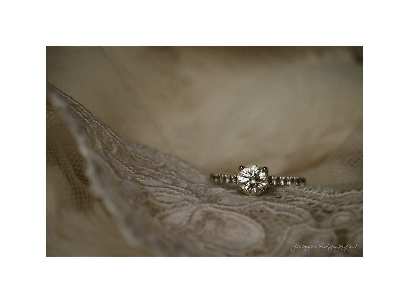 A diamond engagement ring with a solitaire setting is displayed on delicate lace fabric. The background is softly blurred to highlight the sparkle and detail of the ring.