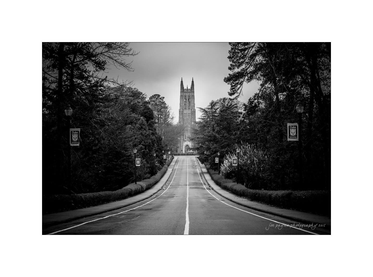 A black and white photograph of an empty road lined with trees, leading to a distant Gothic-style tower. Lamp posts with banners are visible along the road. The sky is overcast, adding a moody atmosphere to the scene.