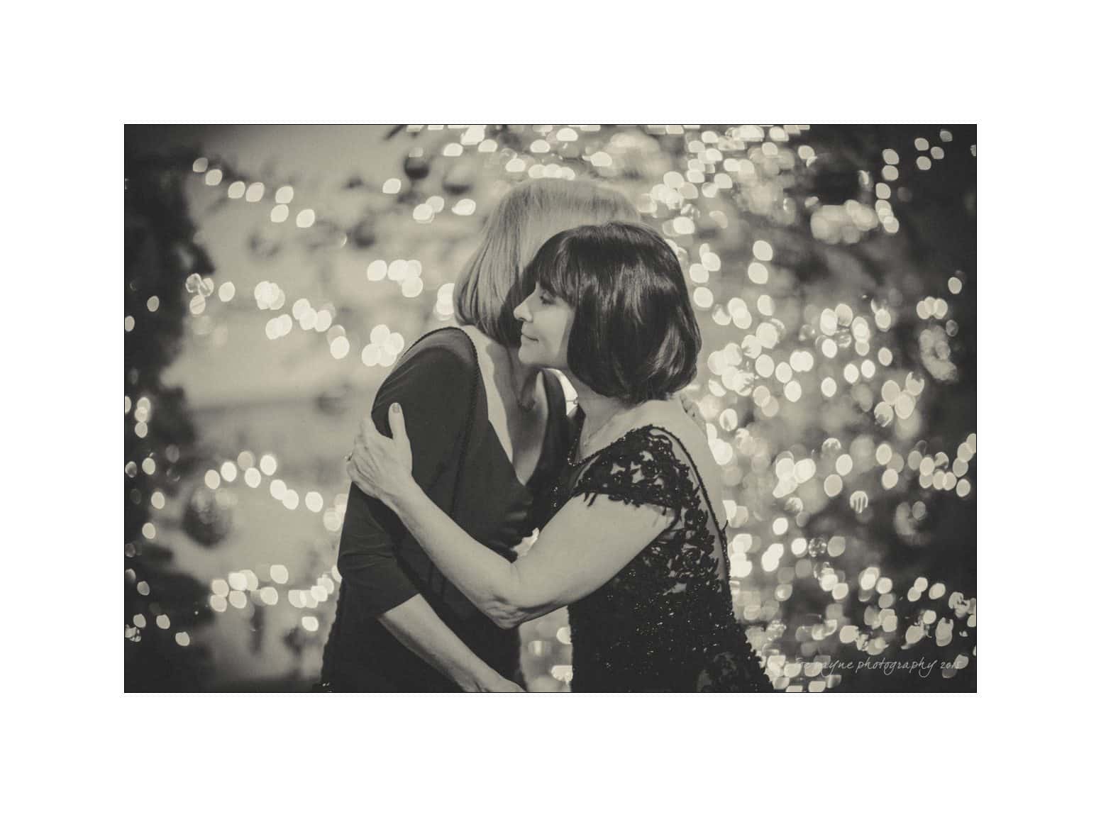 Two women embrace at a festive event in a black and white photograph. One wears a sequined dress, and twinkling lights and a decorated Christmas tree are blurred in the background, creating a warm, celebratory atmosphere.