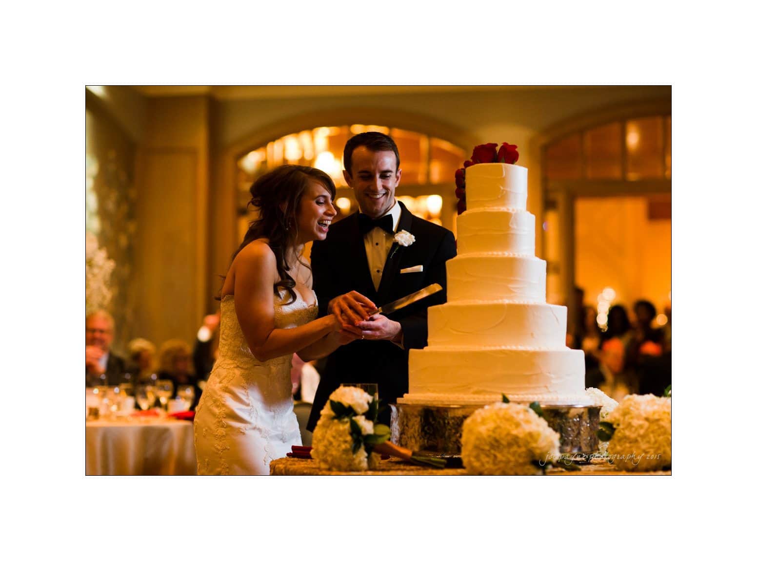 A bride and groom, dressed in a white wedding gown and black tuxedo, smile joyfully as they cut a large, multi-tiered white wedding cake adorned with red roses. The setting is a warmly lit banquet hall with guests in the background.