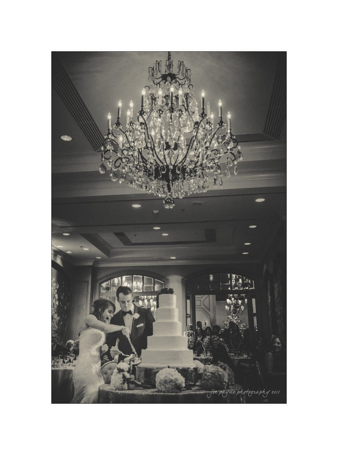 A bride and groom are cutting a tall wedding cake under an elegant chandelier in a dimly lit reception hall. Guests are seated around tables in the background.