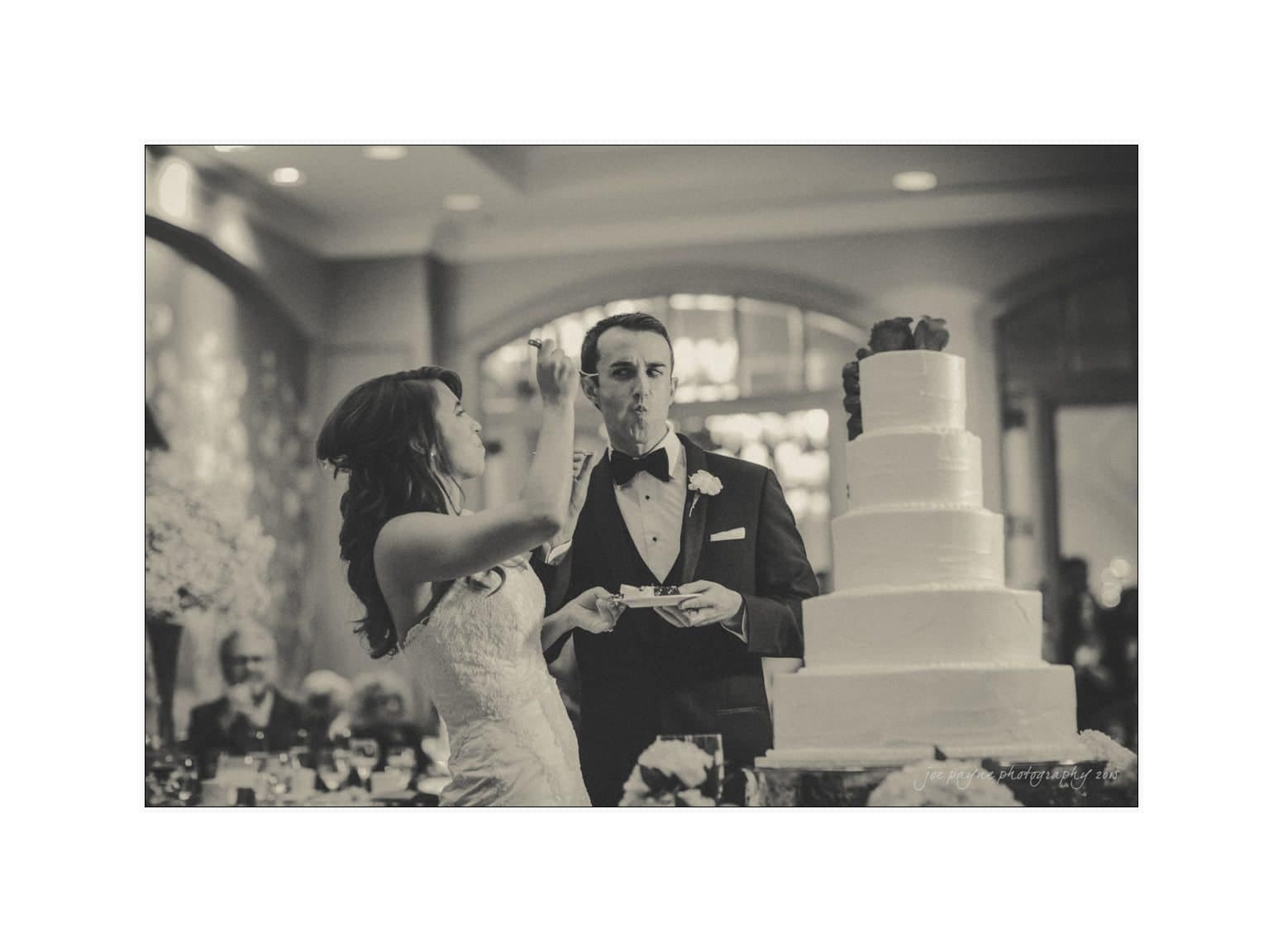 A bride playfully feeds a piece of cake to the groom at their wedding reception. The couple stands beside a tiered cake. The setting is elegant, with guests visible in the softly focused background. The image is in black and white.