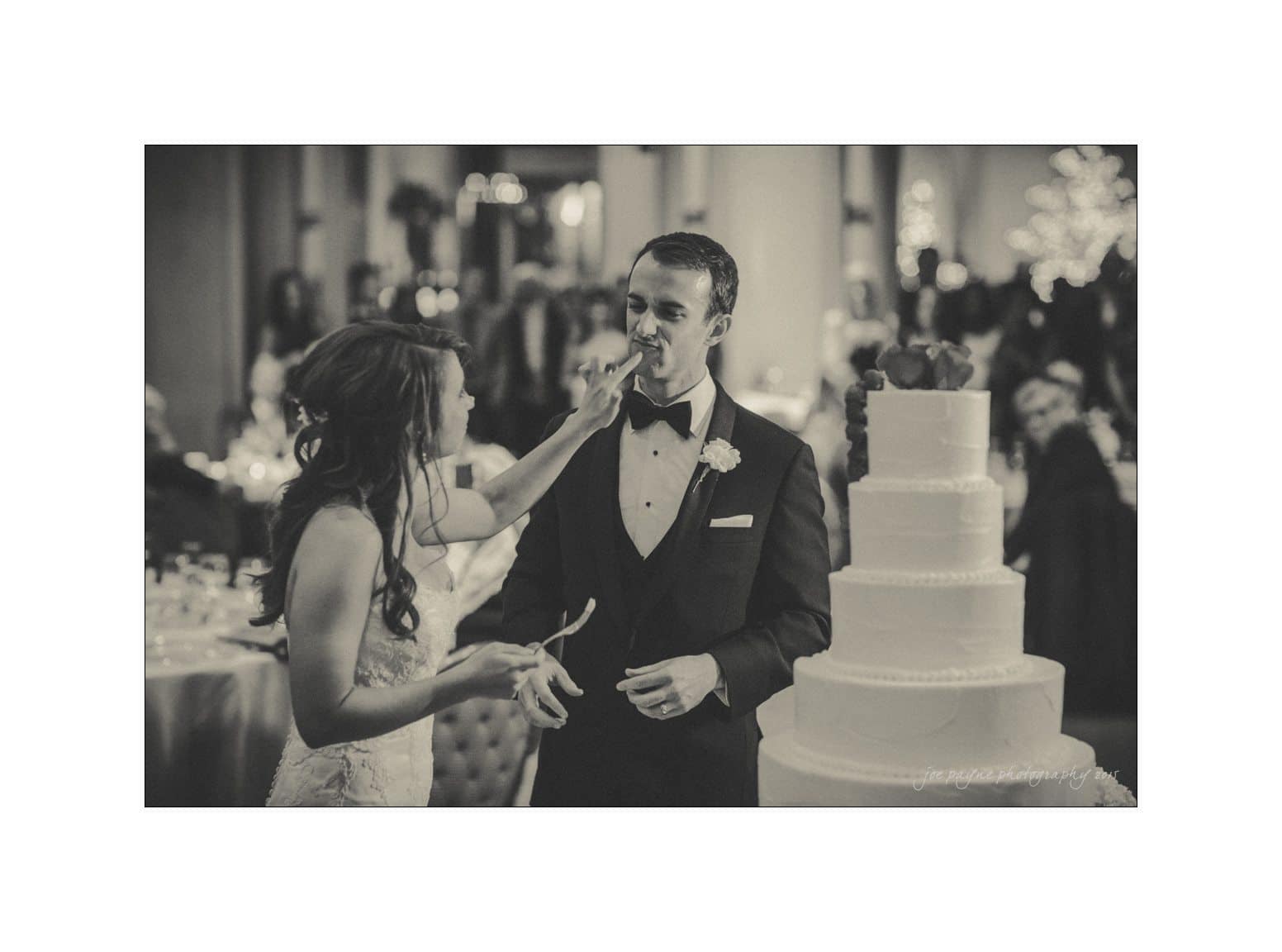 A bride playfully feeds cake to the groom during their wedding reception. They stand beside a multi-tiered wedding cake, surrounded by guests in a warmly lit venue. The image is in black and white, capturing a joyful moment.