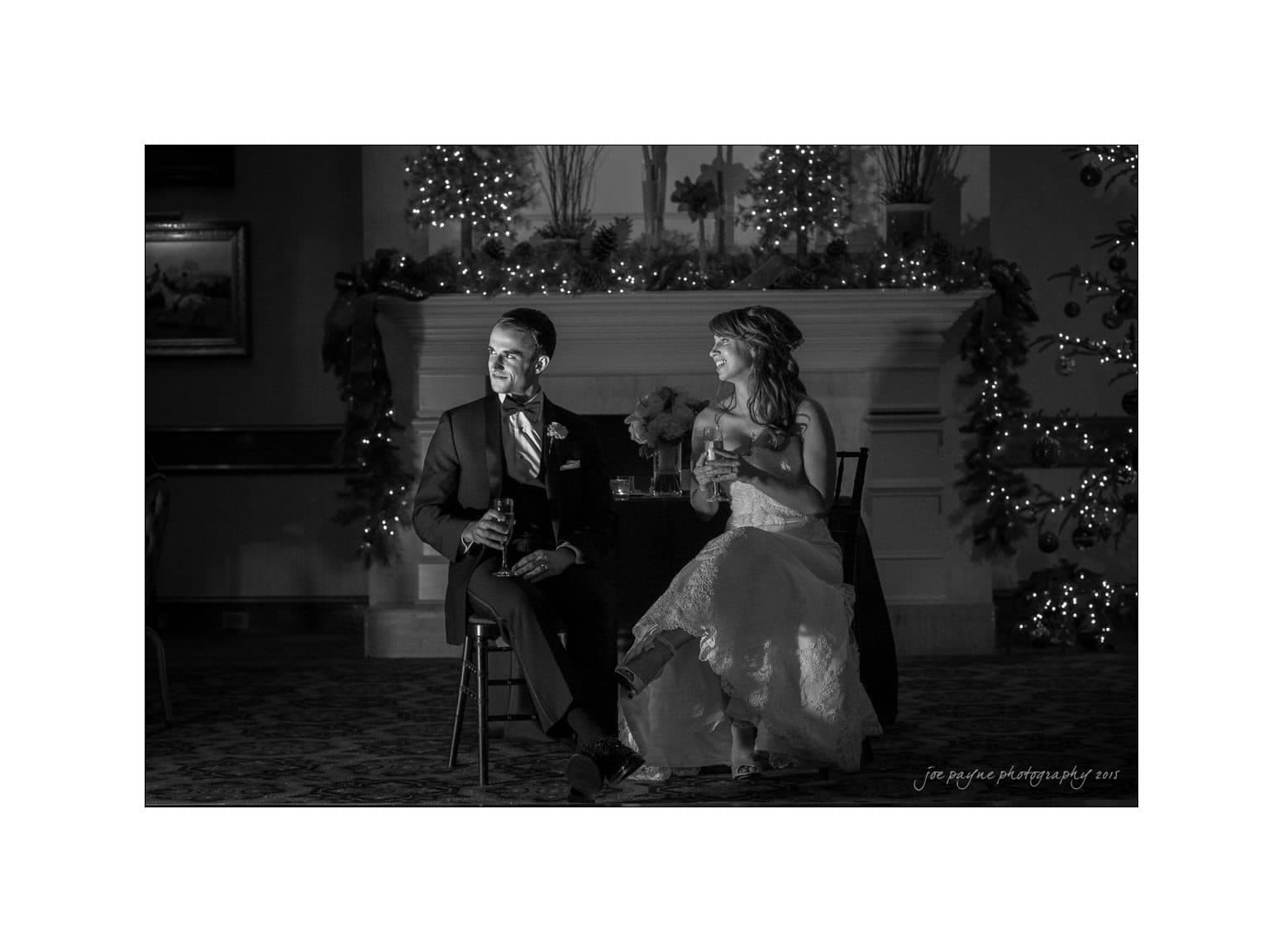 A black and white photo of a bride and groom sitting side by side on chairs, holding drinks. They are smiling, positioned in front of a decorated fireplace adorned with lights and flowers.