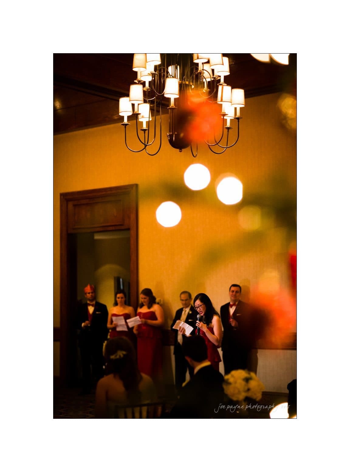 A woman in a dark dress speaks into a microphone at an indoor event, with people in formal attire in the background. A chandelier hangs above, and warm lighting creates a festive atmosphere.