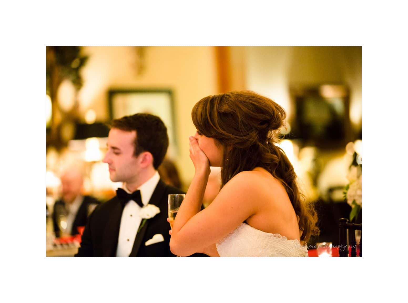 A bride, sitting at a table in a warmly lit room, covers her mouth, possibly laughing or emotional. A groom in a tuxedo is seated next to her, looking on. They are surrounded by soft decor and guests.