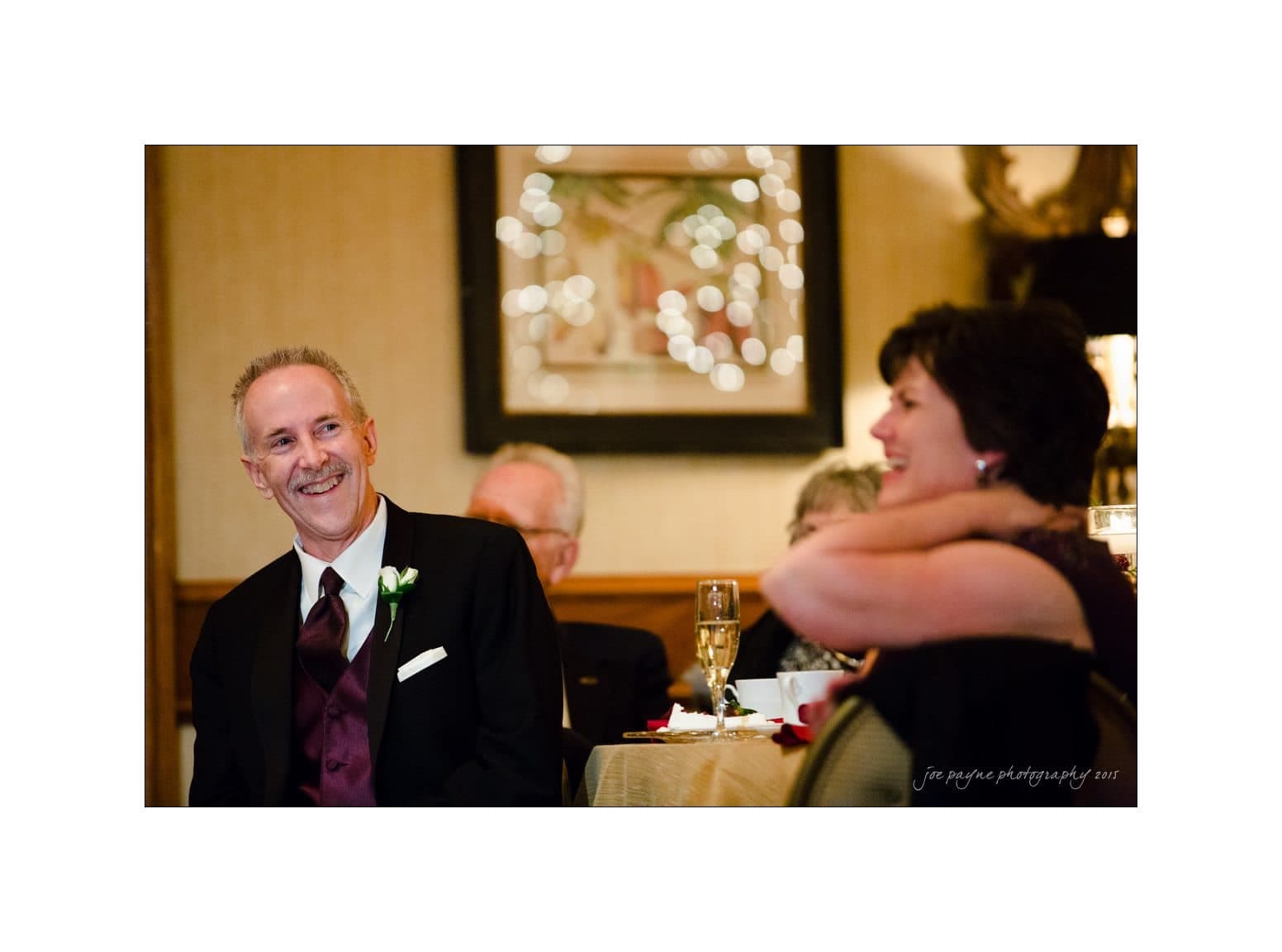 A smiling man in formal attire sits at a table beside a woman, who is also smiling. They are in a warmly lit setting with a decorated wall in the background, suggesting a festive occasion.