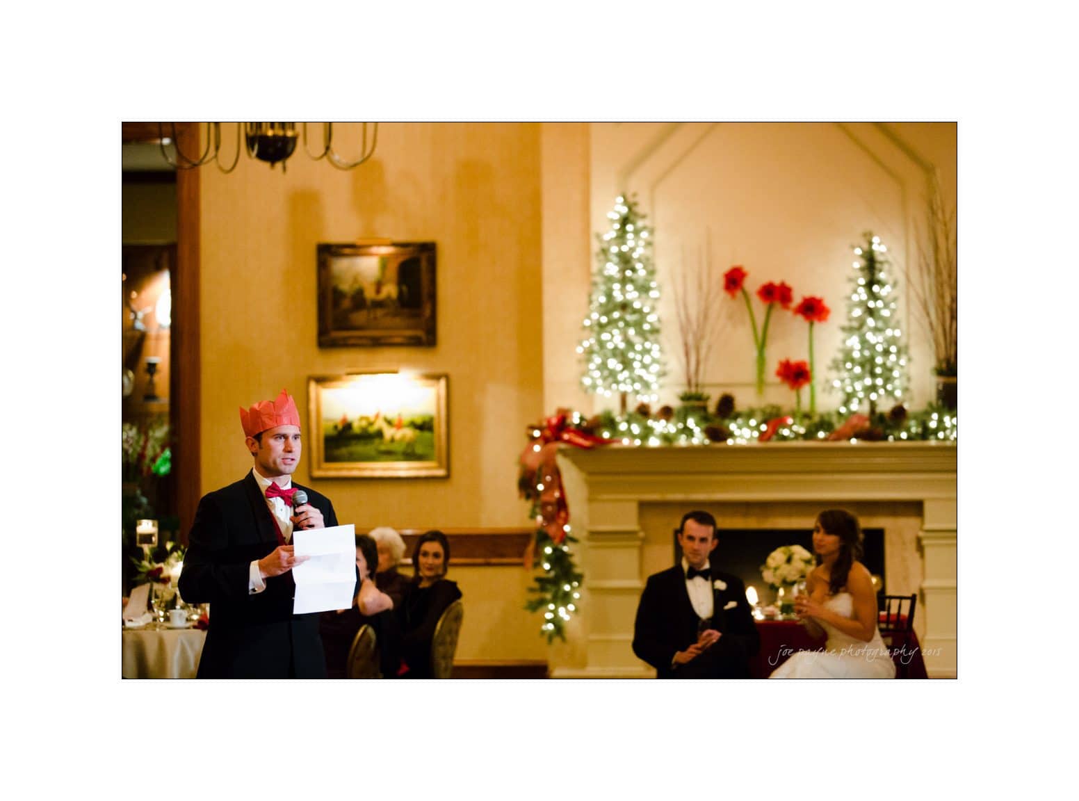 A person wearing a red hat gives a speech at a formal event. A couple, dressed elegantly, sits in the background near a decorated fireplace with lights and large red flowers. Paintings and warm lighting adorn the walls.