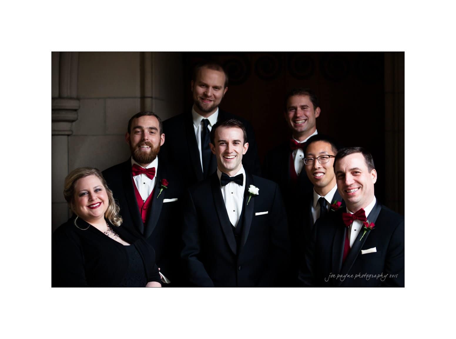 A group of seven people in formal attire, including tuxedos and bow ties, smile at the camera. The individuals are standing closely together in a dimly lit setting, exuding a sense of joy and celebration.