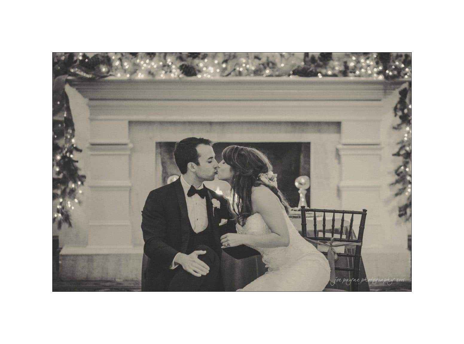 A couple in formal attire shares a kiss in front of a decorated fireplace with string lights. They are seated on chairs, and the image has a vintage, sepia-toned effect.