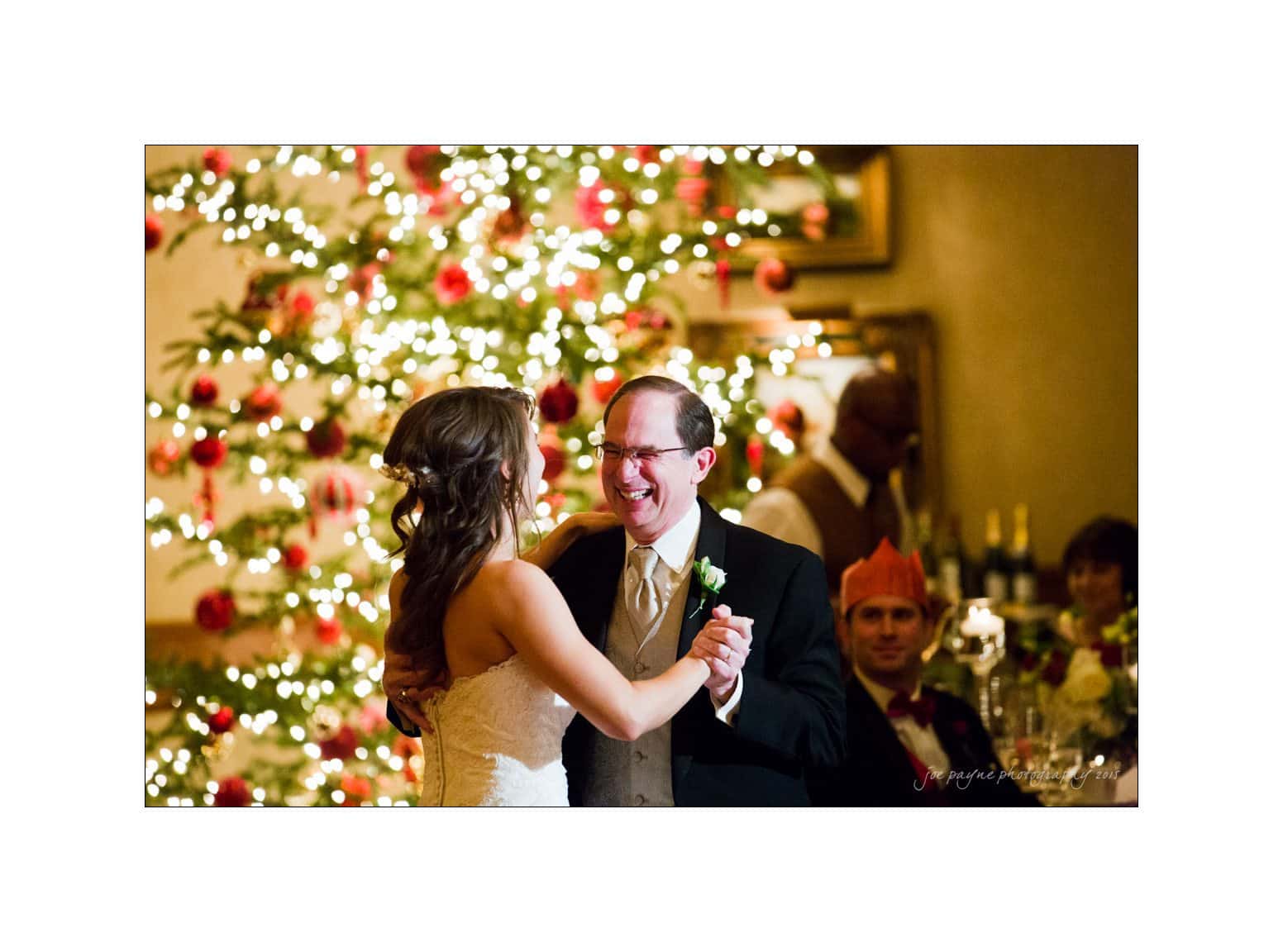 A joyful couple dances together, smiling in front of a brightly lit Christmas tree adorned with red and white ornaments. The groom wears a suit, and the bride is in a white dress. Guests are seated in the dimly lit, festive setting.