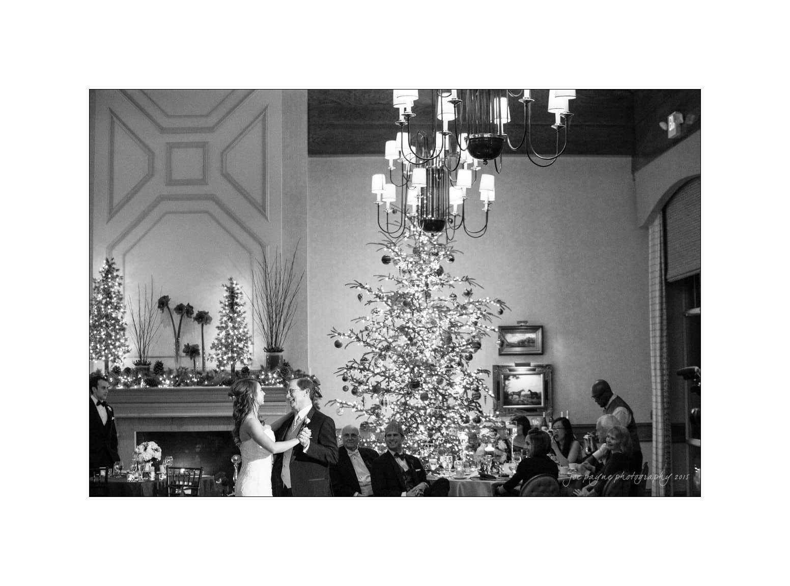 A black and white image of a couple dancing in a festive room. They are surrounded by seated guests at decorated tables. A large, lit Christmas tree and illuminated garlands adorn the background, creating a warm and elegant atmosphere.