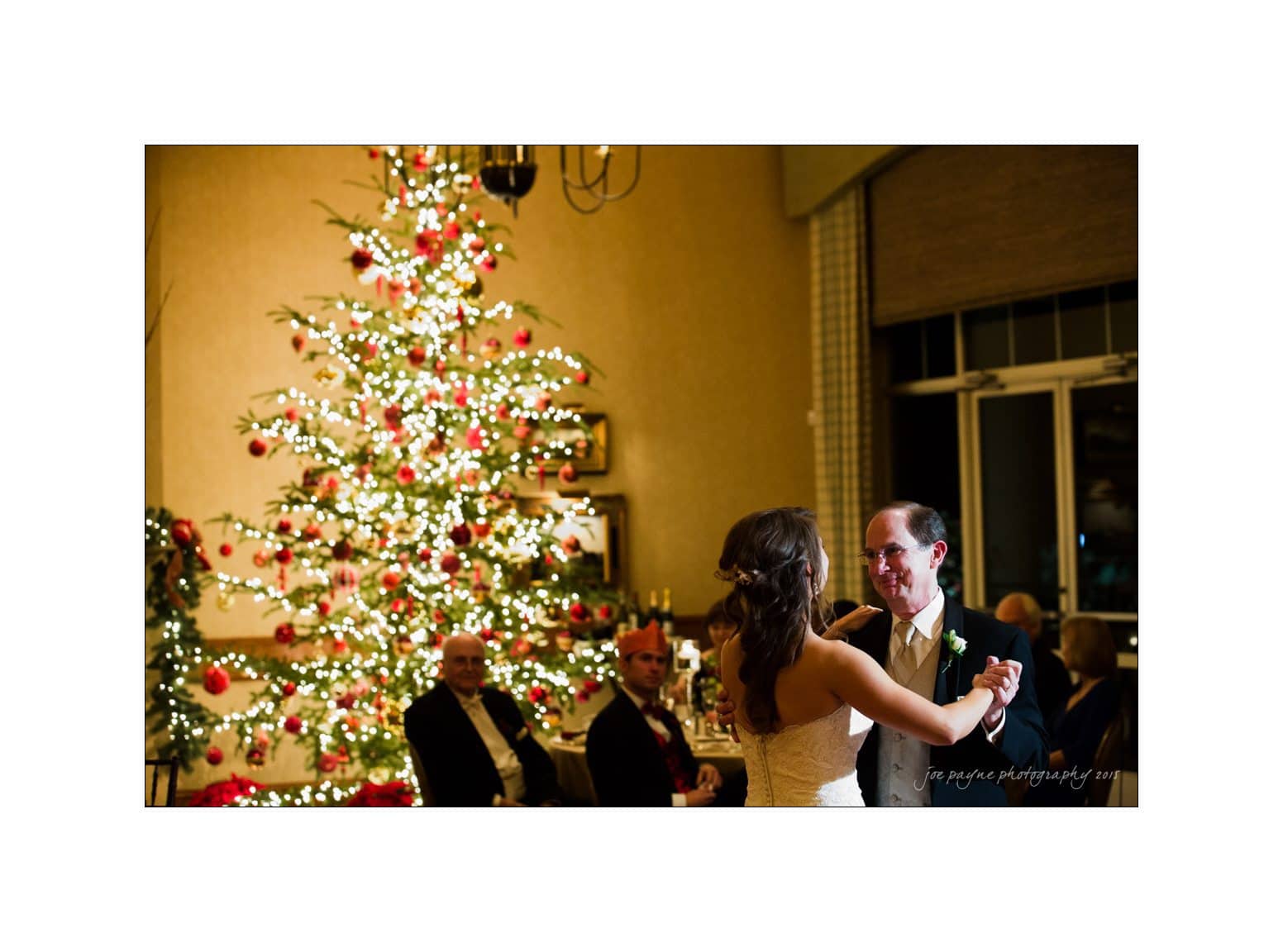A couple shares a dance in a warmly lit room decorated for Christmas. A large, illuminated Christmas tree adorned with red and white ornaments stands in the background, with seated guests watching the dance.