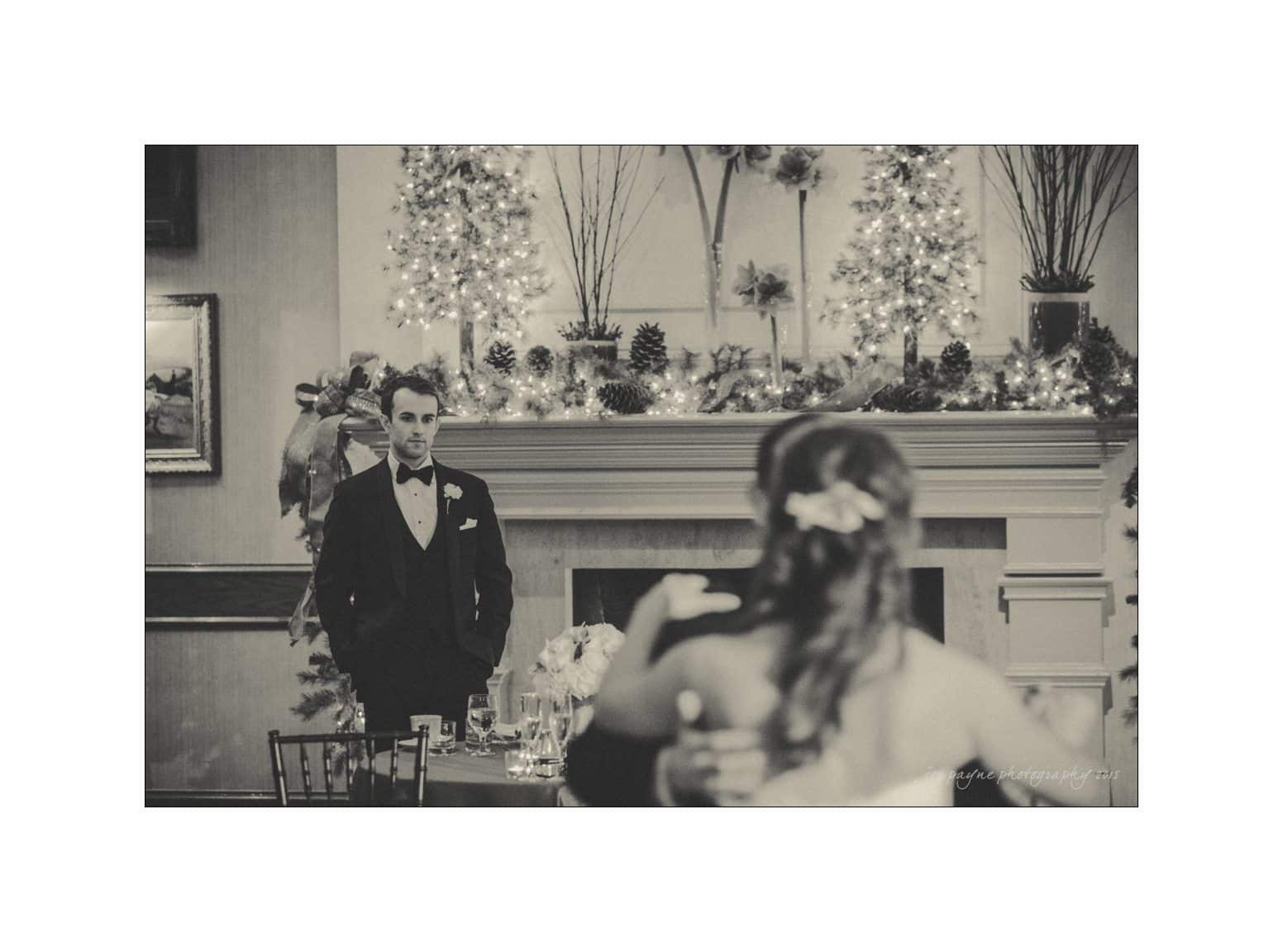 A black and white image of a groom in a suit standing in front of a decorated fireplace with trees and lights. A bride, partially visible from the back, looks toward him. The setting is elegant and festive.