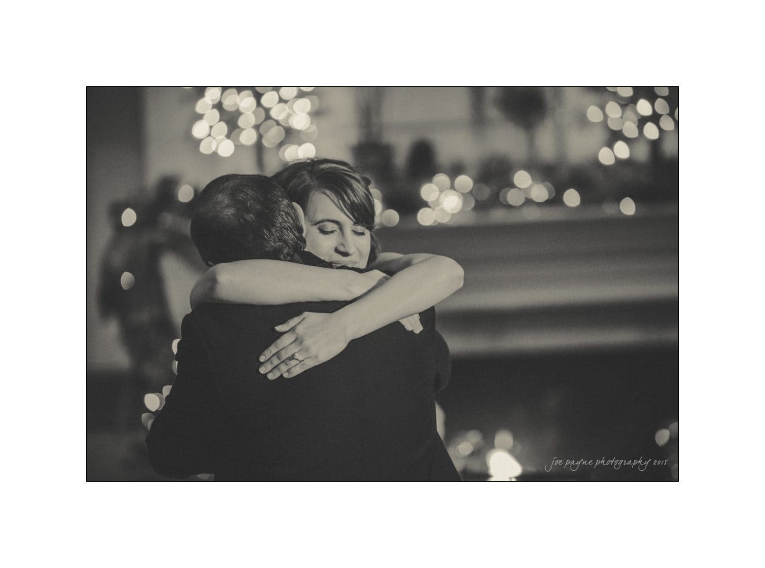 A black and white photo of a woman and a man embracing closely. The background is blurred with twinkling lights, creating a warm, intimate atmosphere.